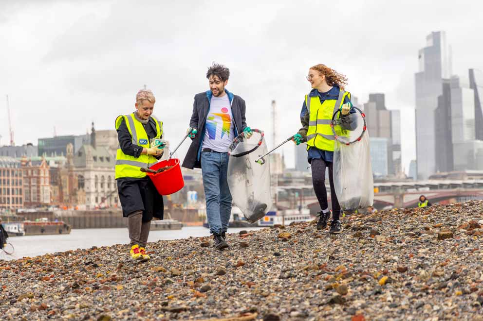Three volunteers cleaning a pebble beach, they are all smiling and there is a city skyline background