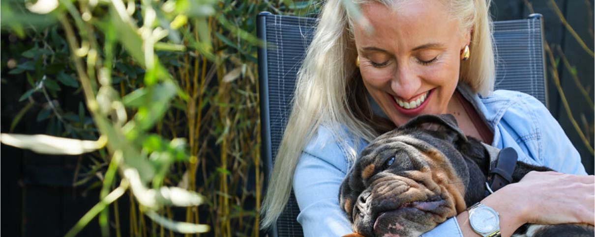 A lady with blonde hair and a blue shirt, smiling down at Bernie, a black and brown bulldog breed dog, she is holding in her arms.