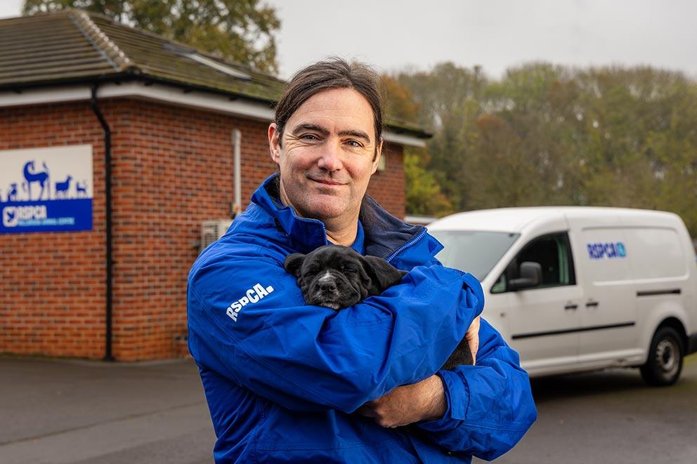 Inspector holds small black puppy, he is standing in front of an RSPCA building and van